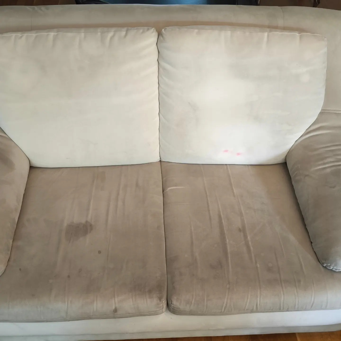 Beżowa kanapa dwuosobowa przed praniem z widocznymi plamami na siedzisku i różowymi plamkami na poduszce oparcia.