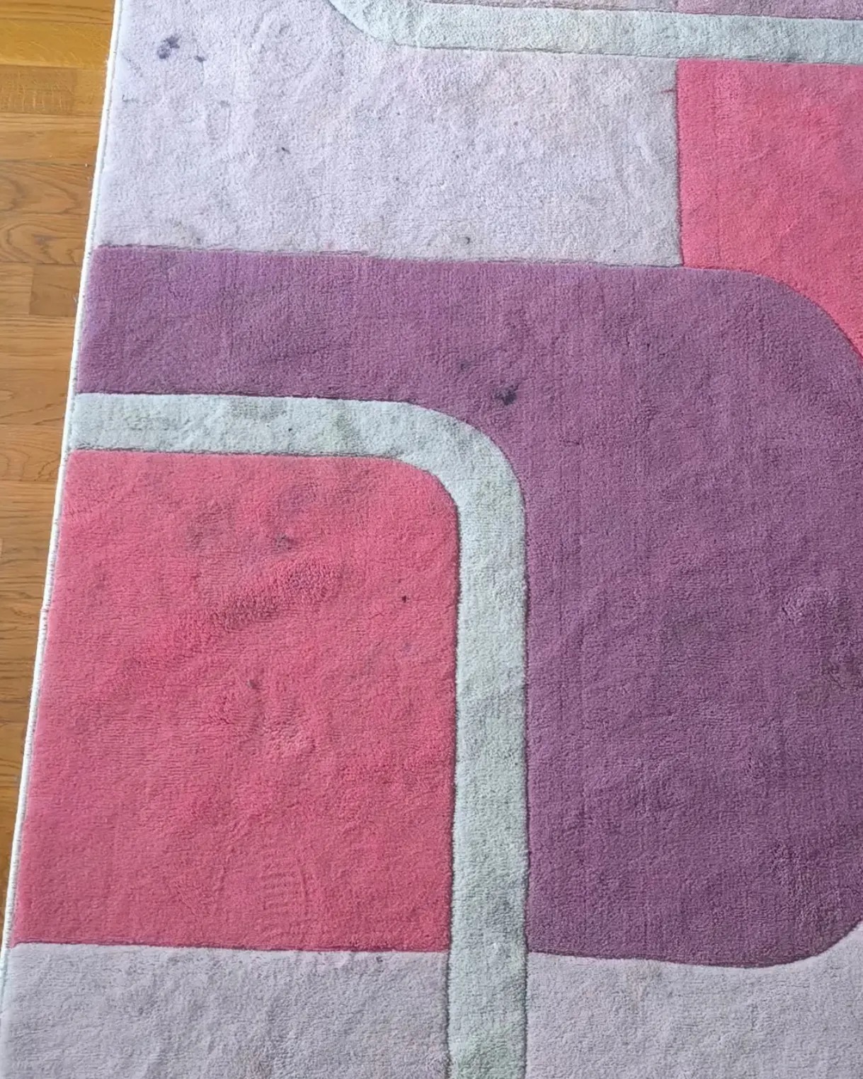 Wielobarwny dywan z geometrycznym wzorem, widoczne plamy i zabrudzenia, leżący na drewnianej podłodze.