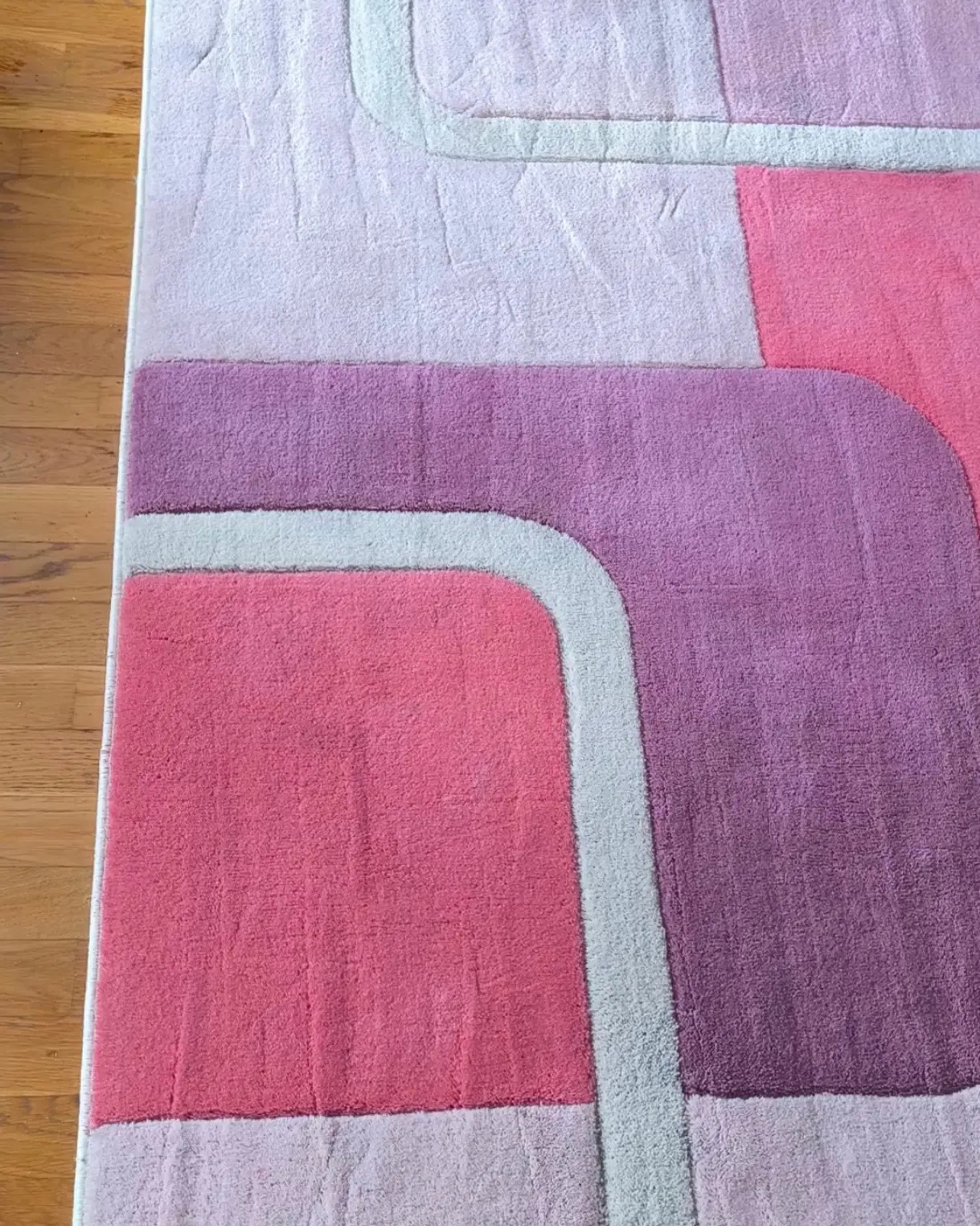 Fragment puszystego dywanu z geometrycznym wzorem w odcieniach różu, fioletu i bieli, leżącego na drewnianej podłodze