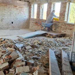 Piotr sierachan - Wnętrze w trakcie rozbiórki: gruz ceglany, pracownik w kombinezonie z taczką przy oknie, odsłonięte cegły, surowy betonowy strop. Widok z bliska na postęp prac.