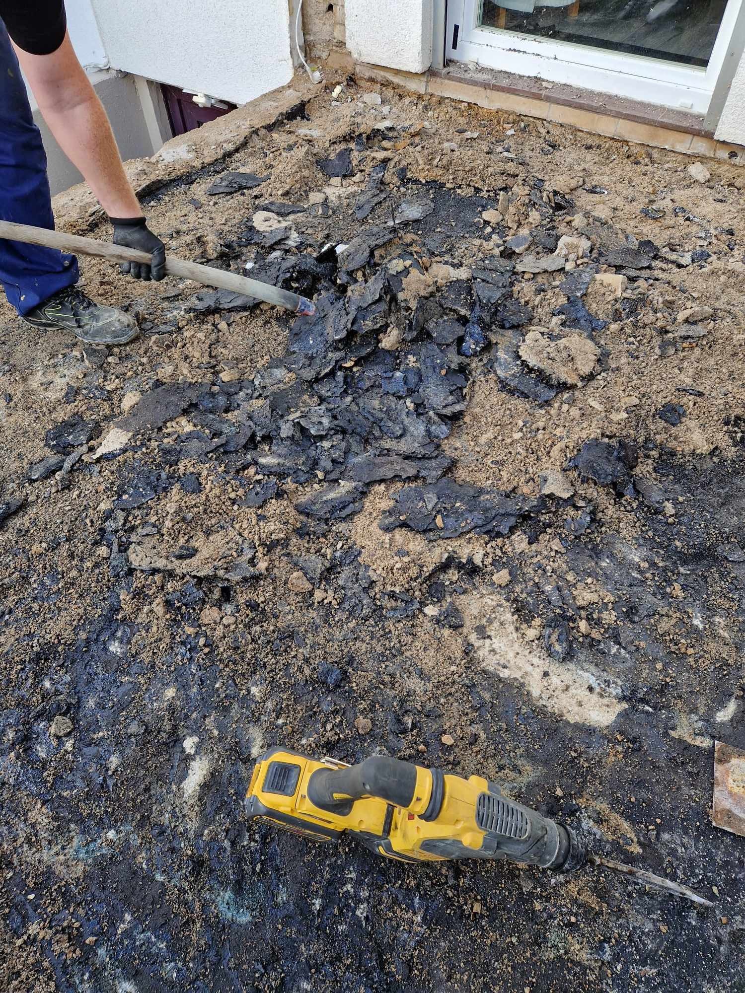 Prace rozbiórkowe przy budynku: robotnik z łopatą usuwa resztki czarnej smoły i gruzu, obok leży żółty młot udarowy.