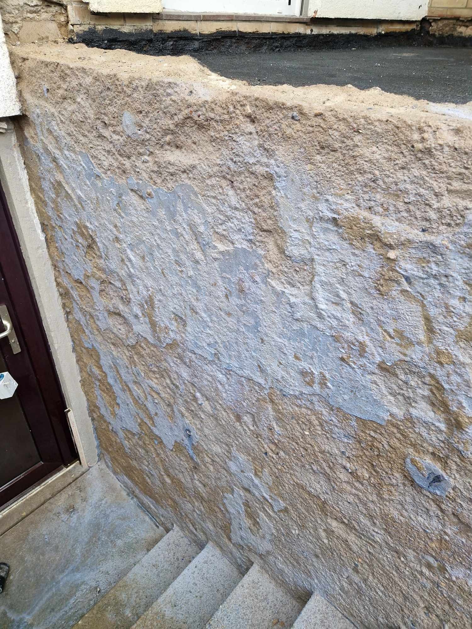 Niedokończona ściana z odsłoniętą strukturą betonu i resztkami starej farby, widoczne schody wejściowe i fragment drzwi.