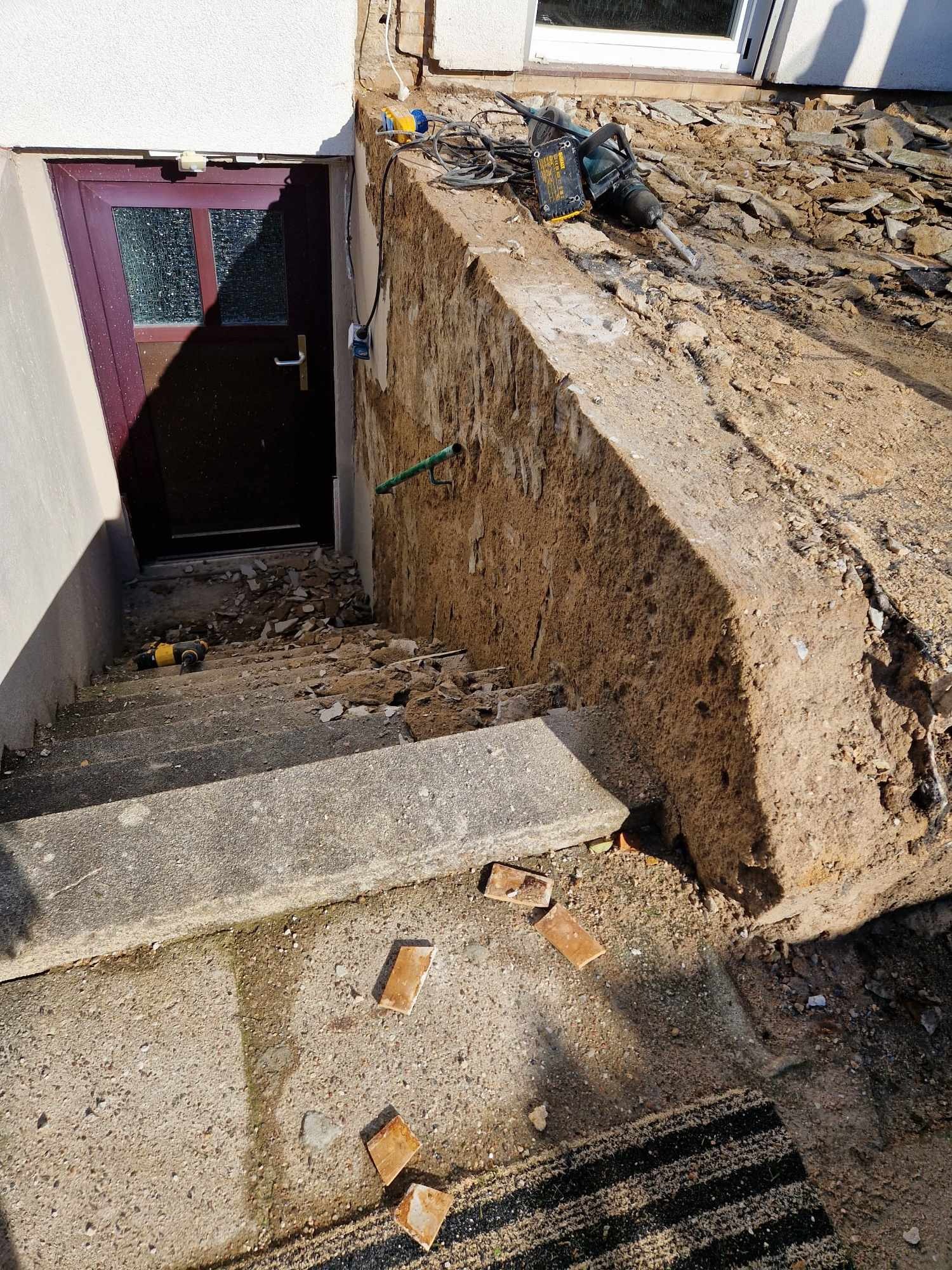 Demontaż betonowej ściany zewnętrznej przy schodach wejściowych do budynku, widoczne narzędzia: młot udarowy i wiertarka, gruz i fragmenty cegieł na schodach.