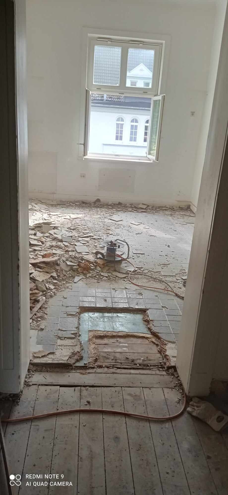 Wnętrze pokoju w trakcie demontażu podłogi, widoczne resztki płytek i odsłonięte deski, zwinięty kabel urządzenia do cięcia płytek leży na podłodze.