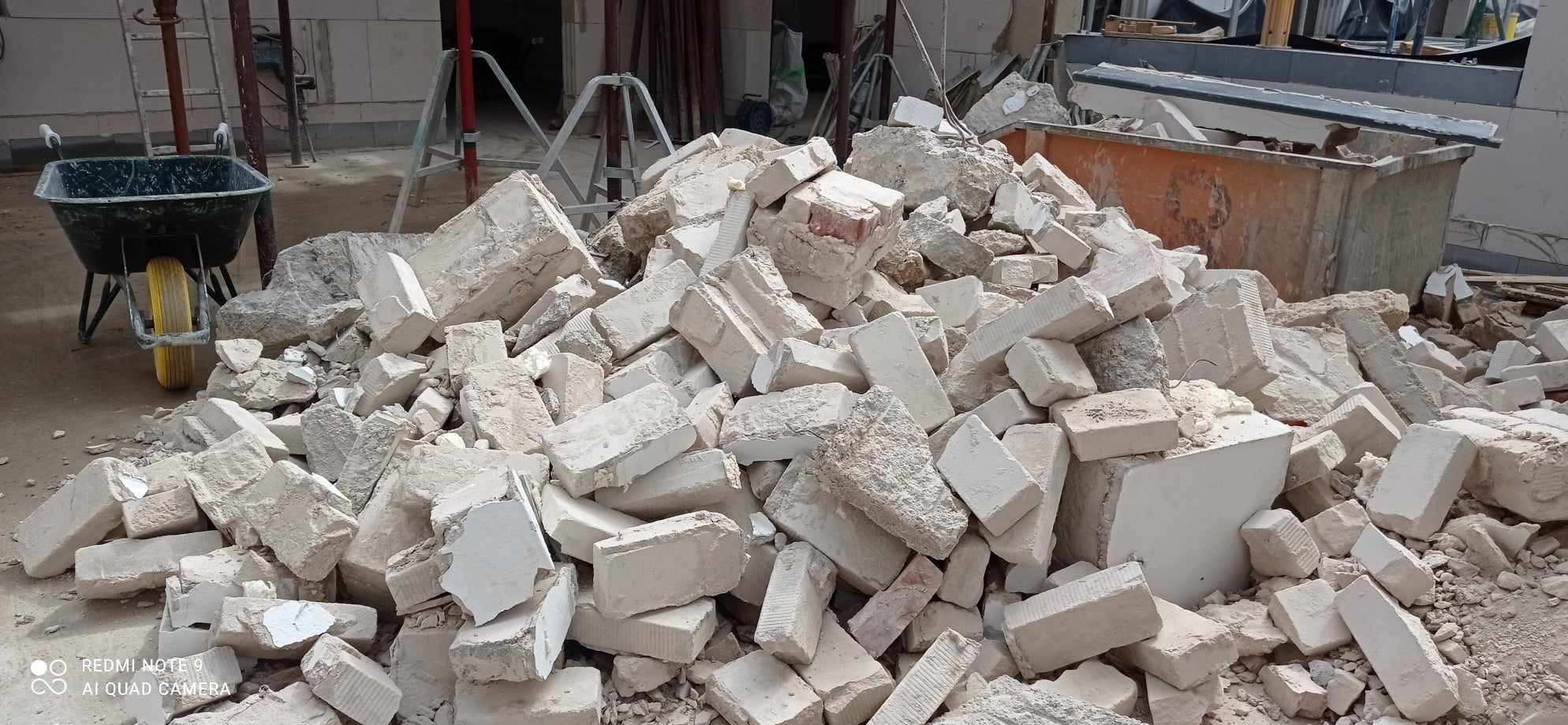 Sterta gruzu z białych bloków po wyburzeniu, widoczna taczka i kontener na odpady budowlane w tle.