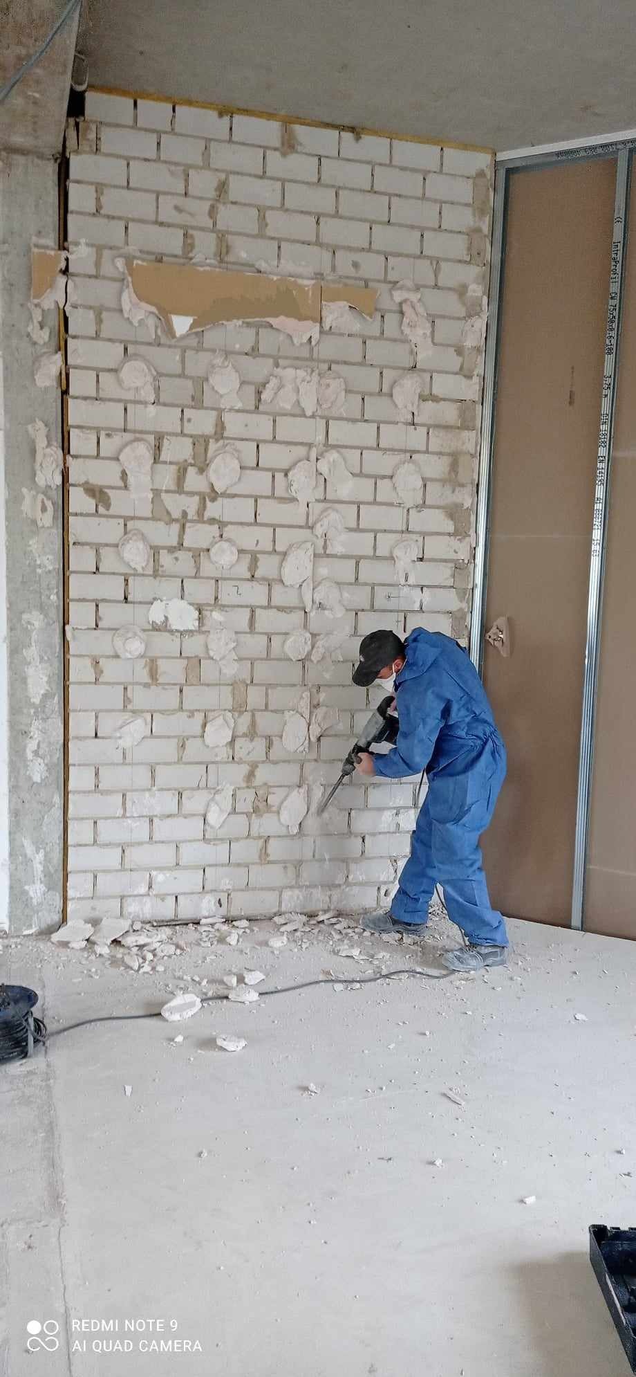 Pracownik w kombinezonie ochronnym z maską i kasku używa młota udarowego do usuwania fragmentów ściany z białej cegły wewnątrz budynku; widoczne metalowe profile konstrukcyjne, gruz na podłodze.