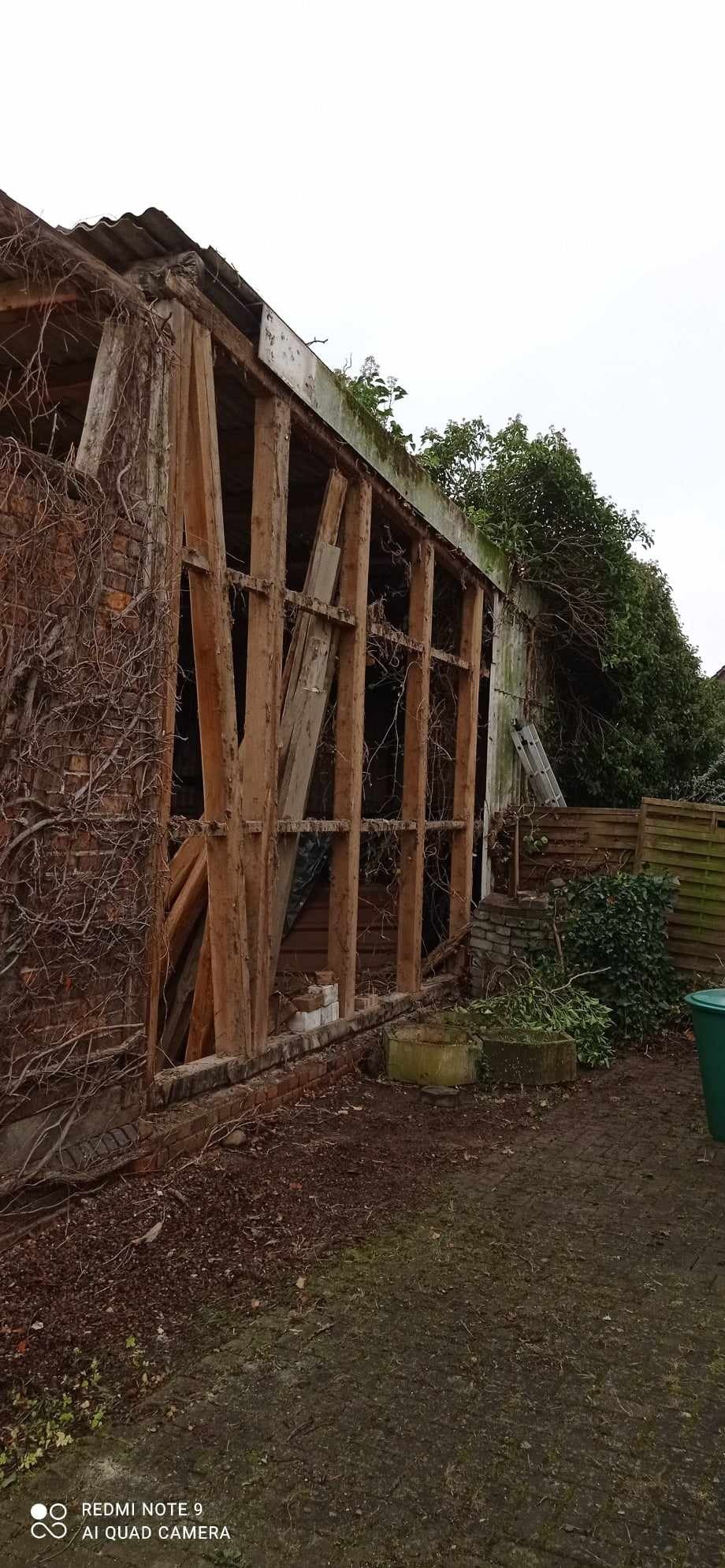 Częściowo rozebrana, ceglana konstrukcja budynku z odsłoniętą drewnianą ramą, porośnięta bluszczem, z widocznymi pozostałościami gruzu i zieleni w otoczeniu.