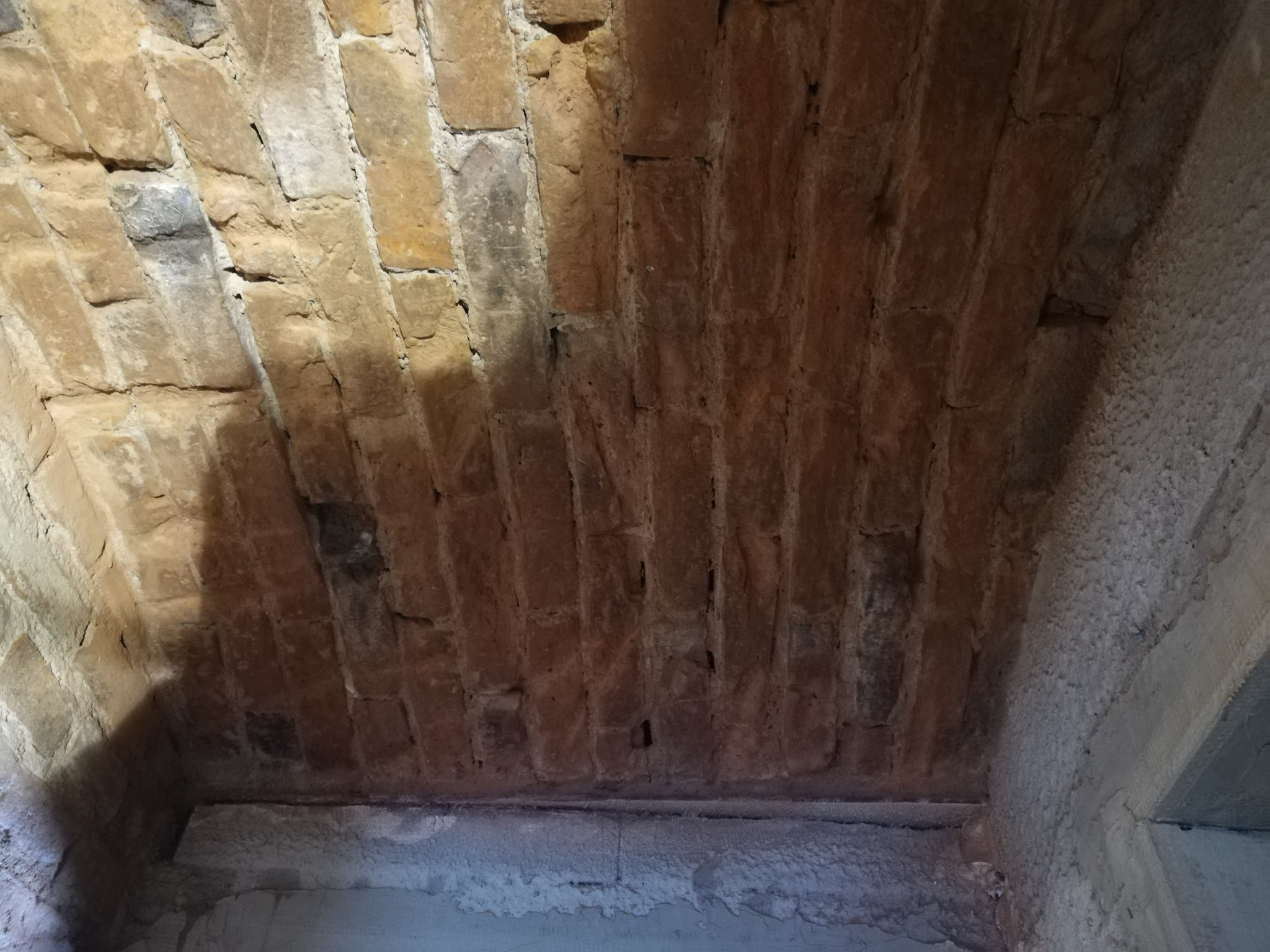 Murowane sklepienie łukowe z cegieł w odcieniach brązu i szarości, widoczne nierówności i ubytki w spoinach, fragment surowej ściany bocznej z betonu.