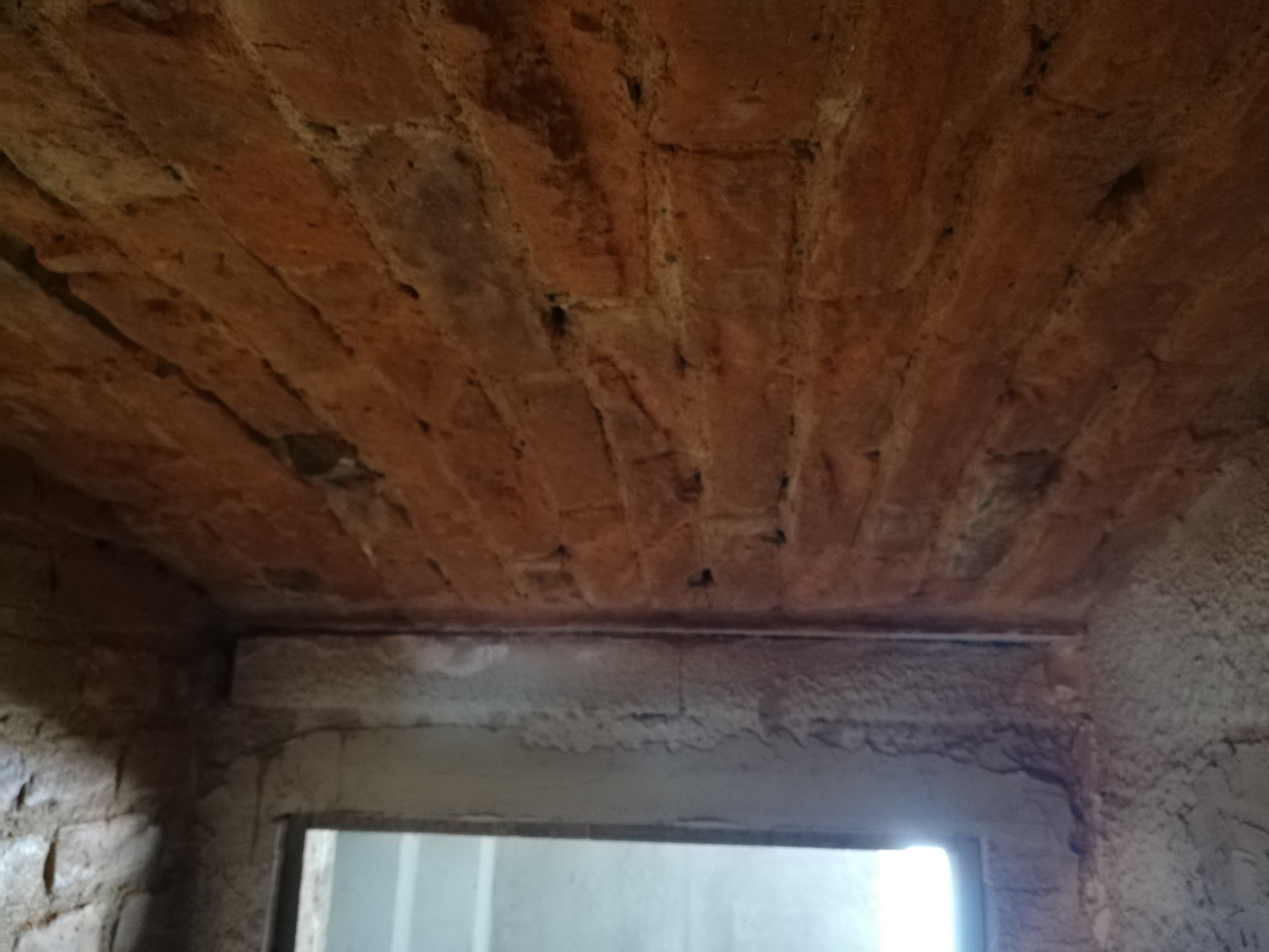 Ceglany łuk sufitowy z widocznymi uszkodzeniami i ubytkami, fragment ściany z surowym tynkiem oraz otwór drzwiowy z widocznym światłem w tle.