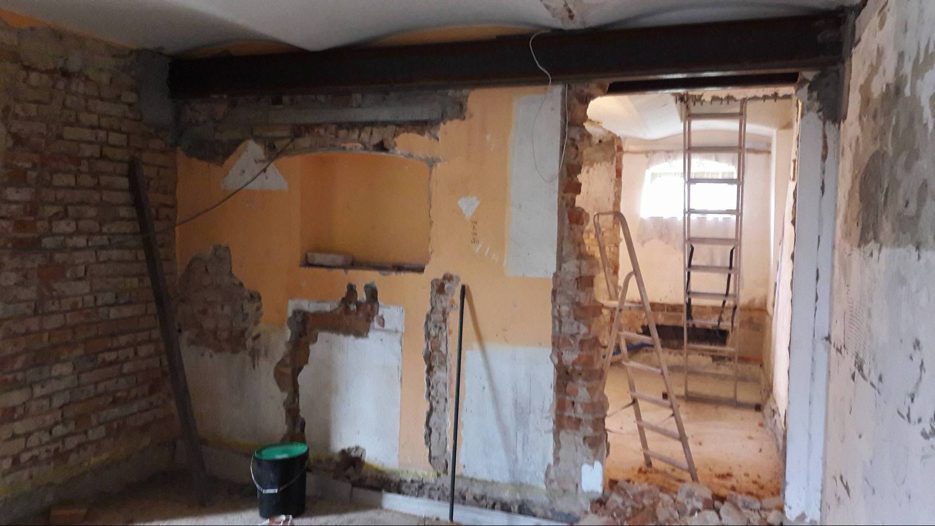 Wnętrze w trakcie demontażu ścian, widoczne fragmenty cegieł, instalacje i drabina w przejściu do kolejnego pomieszczenia.
