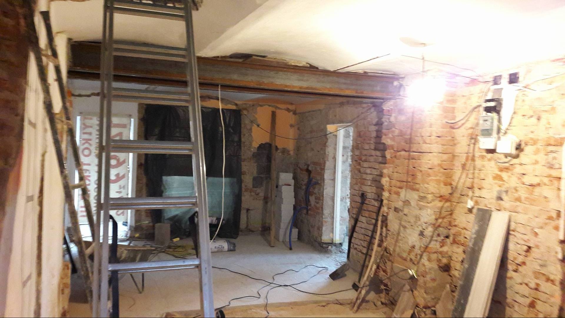 Wnętrze w trakcie prac remontowych: odsłonięta cegła, metalowa belka wzmacniająca strop, aluminiowa drabina, przewody elektryczne i gruz.