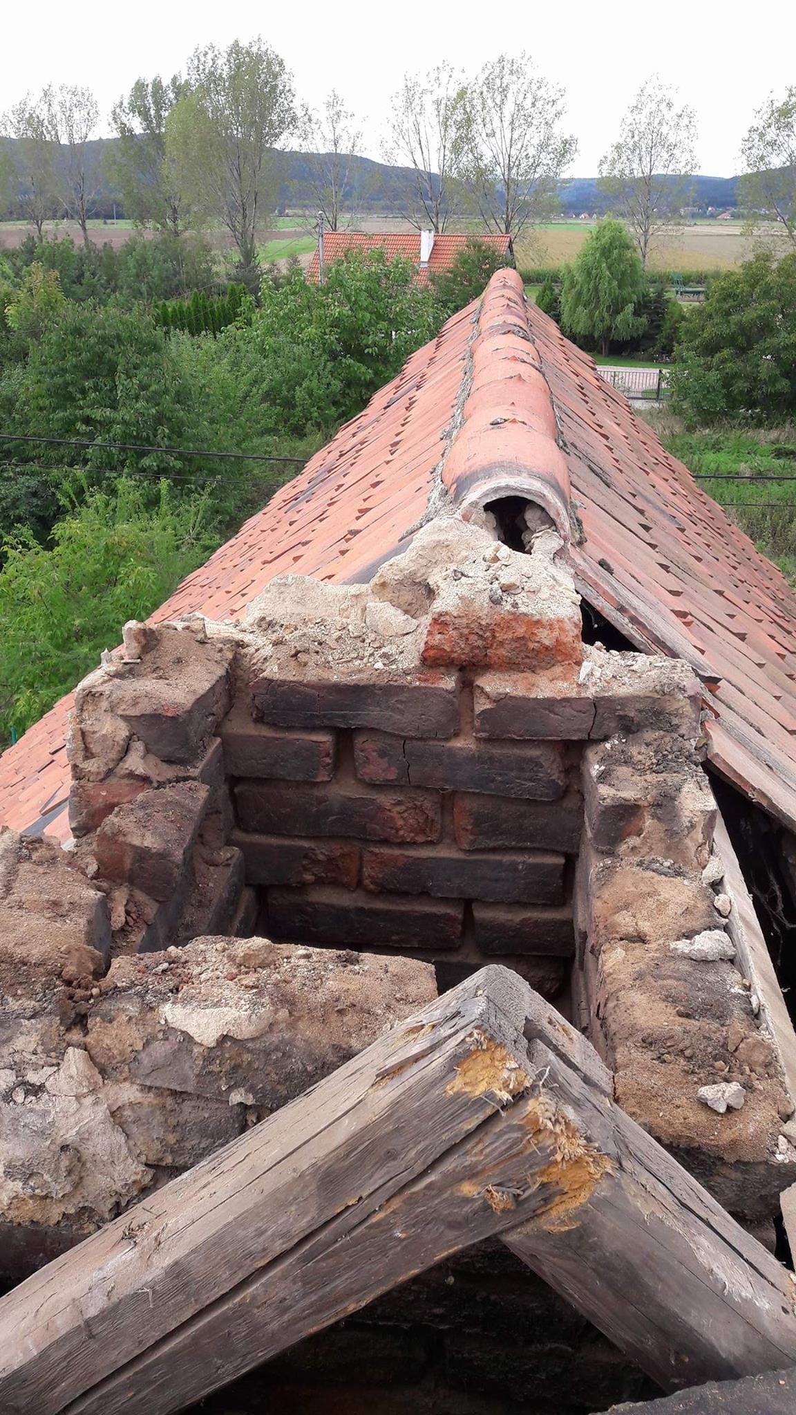 Uszkodzony komin z cegły na dachu z czerwonej dachówki, widoczne elementy konstrukcji drewnianej, w tle zieleń i zabudowania.