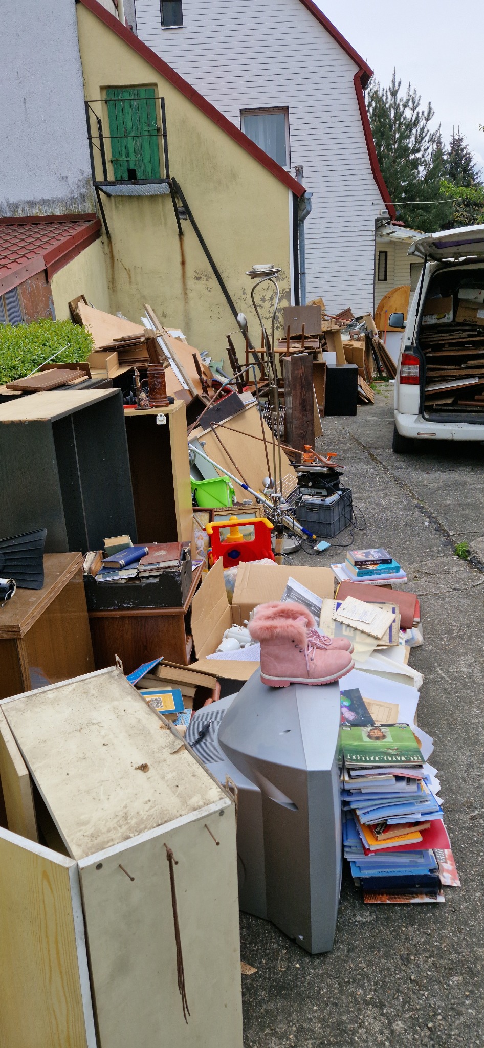 Sterta mebli, książek i sprzętów domowych przygotowana do wywiezienia, z różowym butem na telewizorze kineskopowym. Część rzeczy załadowana do białego busa.
