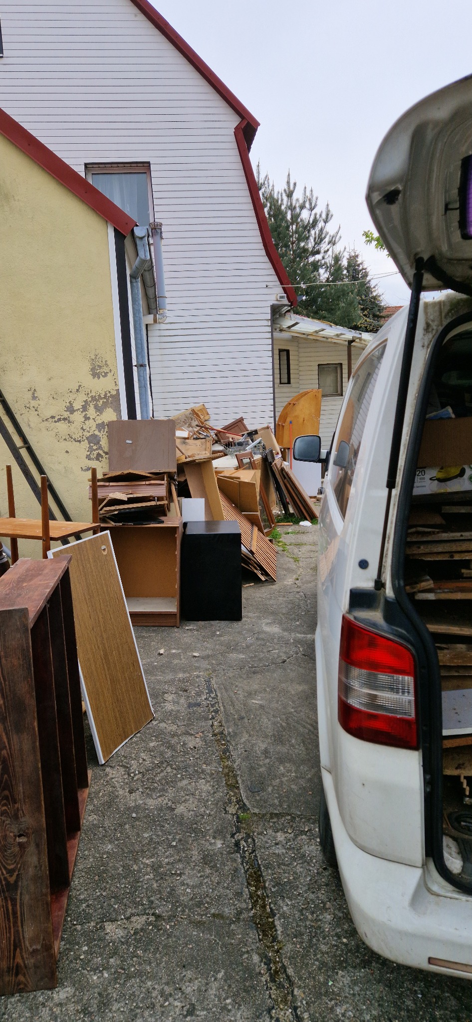 Sterta gruzu i elementów mebli przygotowana do wywozu, częściowo załadowana do bagażnika białego samochodu dostawczego, w tle budynki mieszkalne.