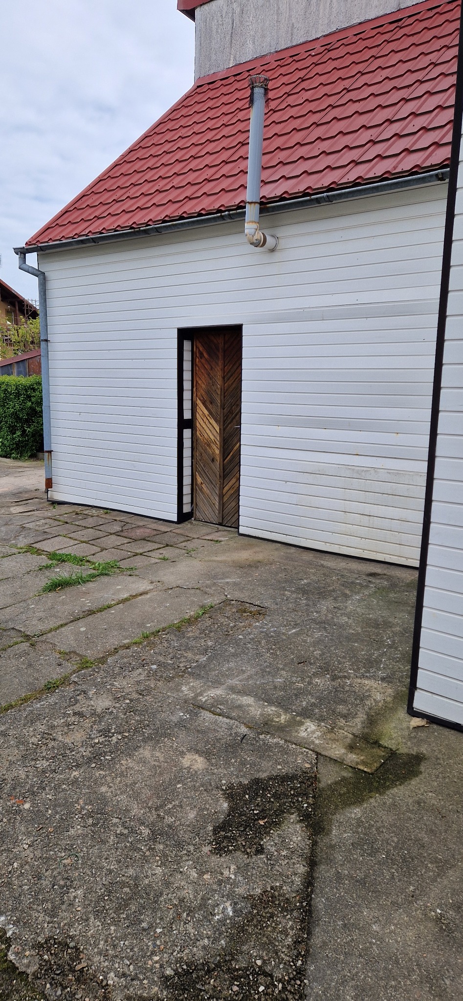 Betonowy budynek gospodarczy z drewnianymi drzwiami i czerwoną dachówką, widok z zewnątrz na betonowy podjazd.