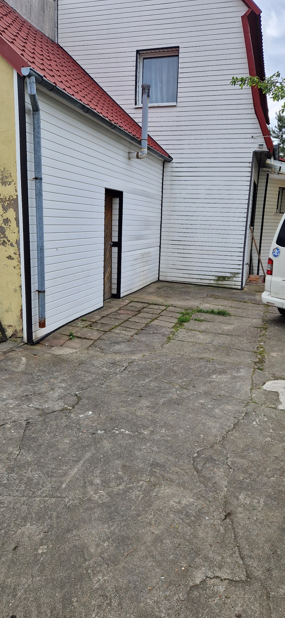 Fragment posesji z budynkiem o białej elewacji z sidingu, czerwonym dachem i betonową nawierzchnią z widocznymi pęknięciami i porostami.