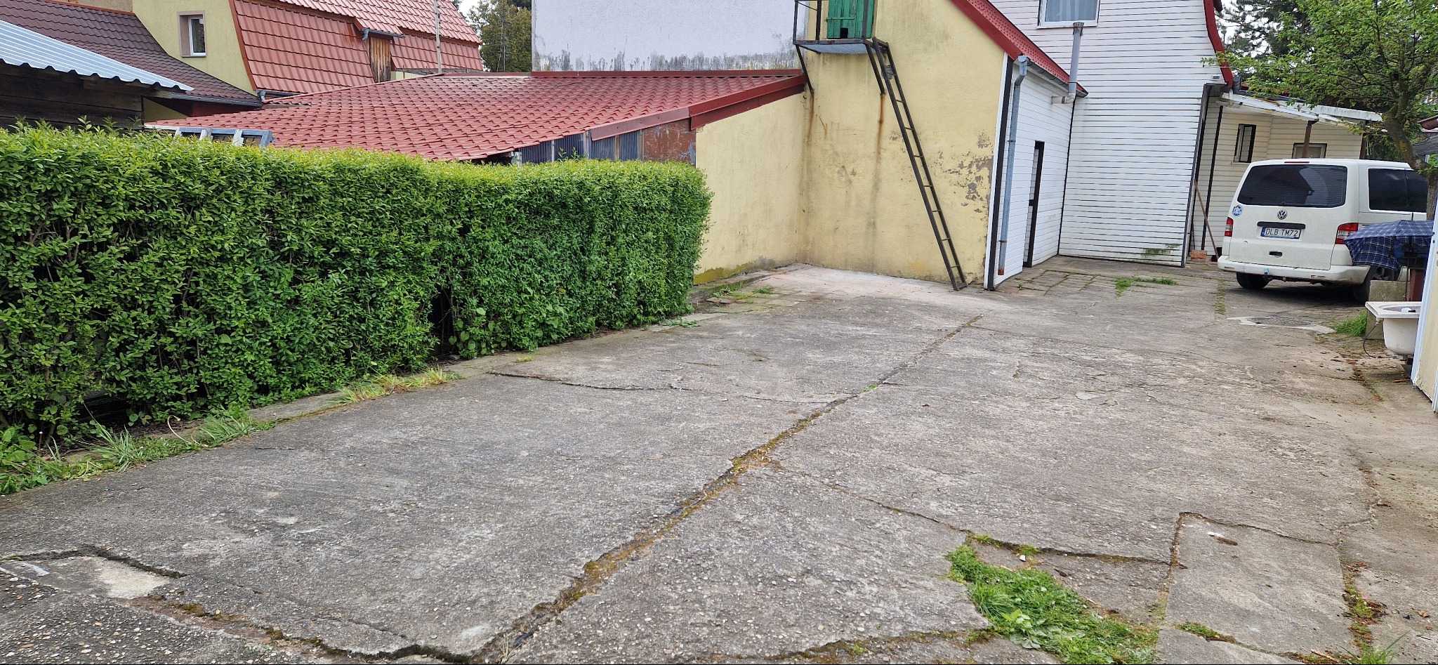 Podjazd z popękanego betonu, obok gęsty żywopłot i budynki gospodarcze z metalową drabiną, w tle biały bus.