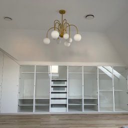 Meble Ledwoń Dawid Ledwoń - Biała szafa wnękowa z przeszklonymi drzwiami w poddaszu, oświetlona złotym żyrandolem z kulistymi kloszami, widoczne skosy i okno dachowe.
