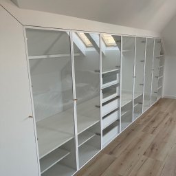 Meble Ledwoń Dawid Ledwoń - Biała zabudowa garderoby z przeszklonymi frontami, dopasowana do skosu poddasza, z widocznymi półkami i szufladami, na podłodze panele imitujące drewno.