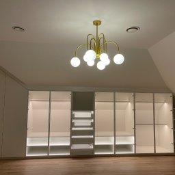 Meble Ledwoń Dawid Ledwoń - Biała szafa wnękowa z oświetleniem LED wewnątrz, na poddaszu, z drewnianą podłogą i złotym, nowoczesnym żyrandolem.