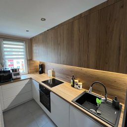 Meble Ledwoń Dawid Ledwoń - Nowoczesna kuchnia z drewnianymi szafkami górnymi i białymi dolnymi, blatem imitującym drewno oraz czarnym zlewem z baterią i dozownikiem na mydło. Na blacie stoją przyprawy i butelka z oliwą.