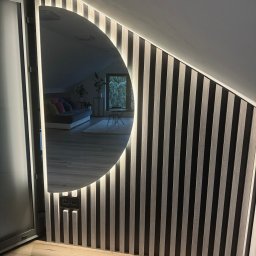 Meble Ledwoń Dawid Ledwoń - Ściana z drewnianych lameli z wbudowanym, podświetlanym lustrem w kształcie półksiężyca, odbijającym wnętrze pokoju na poddaszu z szarą sofą i roślinami.