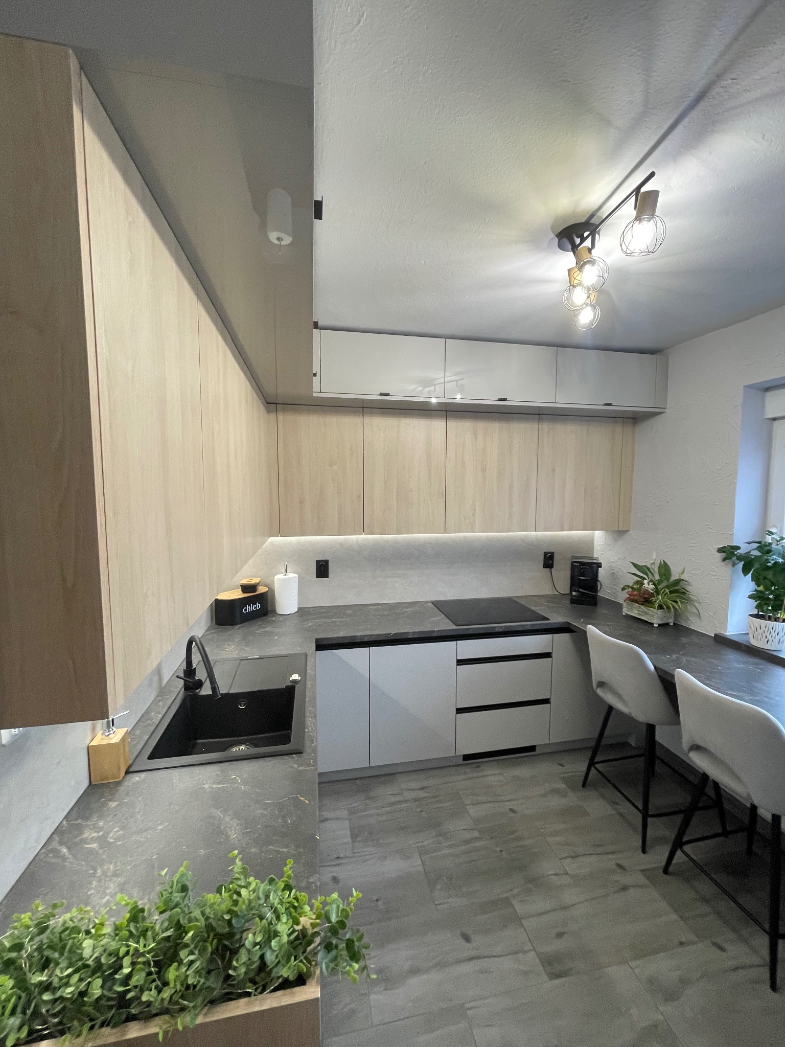 Nowoczesna kuchnia z drewnianymi frontami szafek, czarnym zlewem i blatem z ciemnego kamienia, oświetlona lampą sufitową z żarówkami w stylu Edison.