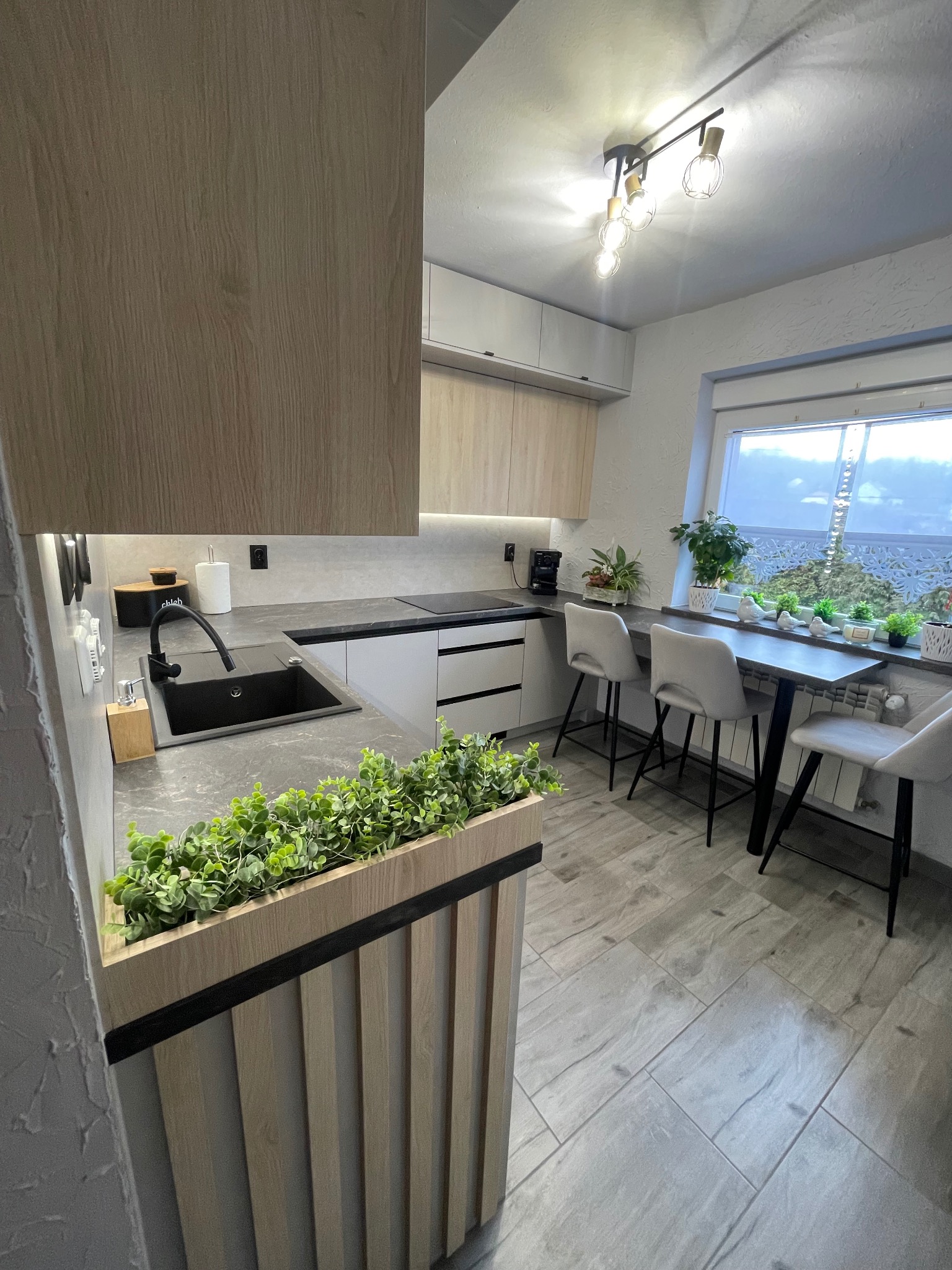 Jasna kuchnia z drewnianymi frontami, ciemnym blatem i czarnym zlewem, ozdobiona zielenią w doniczkach na blacie oraz w dekoracyjnej skrzynce na wyspie kuchennej, z trzema krzesłami barowymi przy...