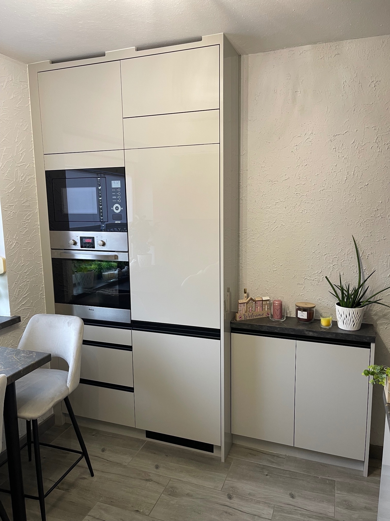 Biała, nowoczesna zabudowa kuchenna z piekarnikiem i mikrofalówką, uzupełniona dekoracjami na blacie i krzesłem barowym z szarą tapicerką.