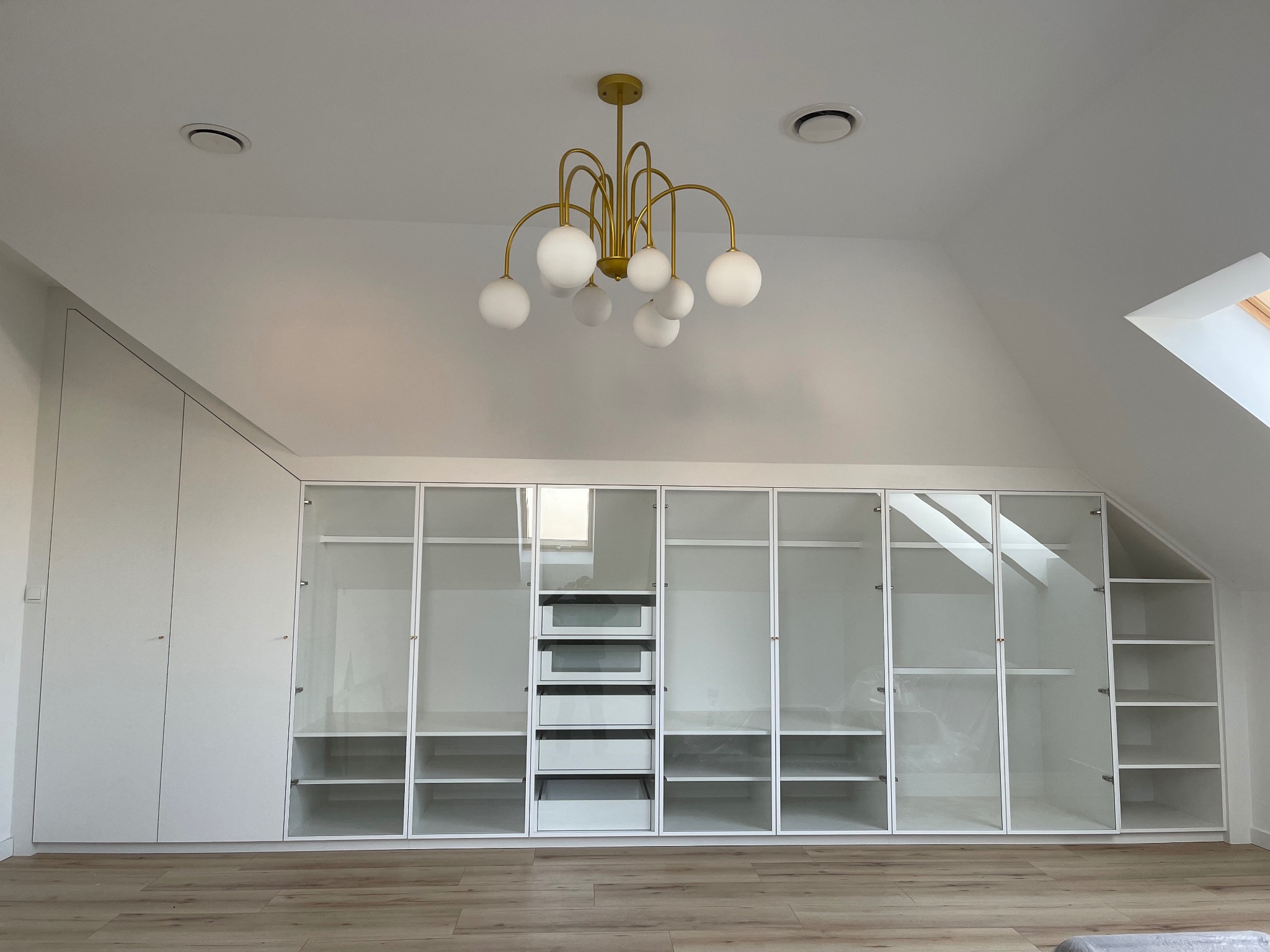 Biała szafa wnękowa z przeszklonymi drzwiami w poddaszu, oświetlona złotym żyrandolem z kulistymi kloszami, widoczne skosy i okno dachowe.