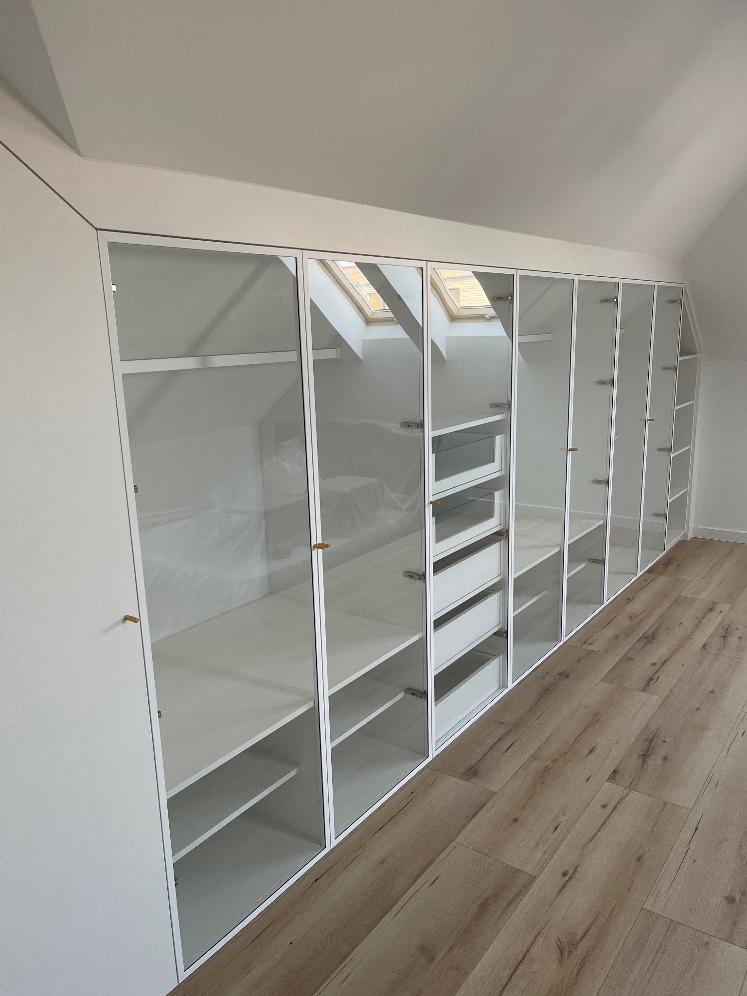 Biała zabudowa garderoby z przeszklonymi frontami, dopasowana do skosu poddasza, z widocznymi półkami i szufladami, na podłodze panele imitujące drewno.