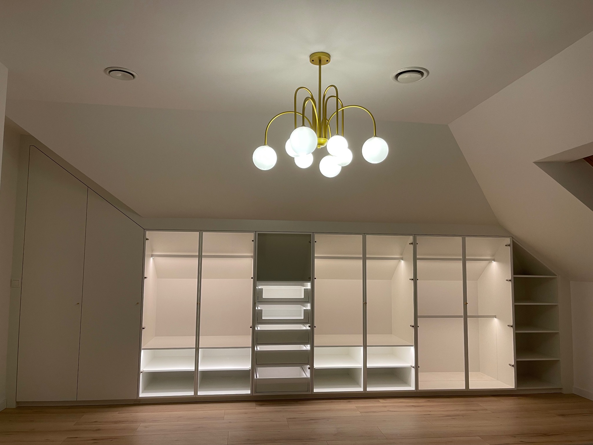 Biała szafa wnękowa z oświetleniem LED wewnątrz, na poddaszu, z drewnianą podłogą i złotym, nowoczesnym żyrandolem.