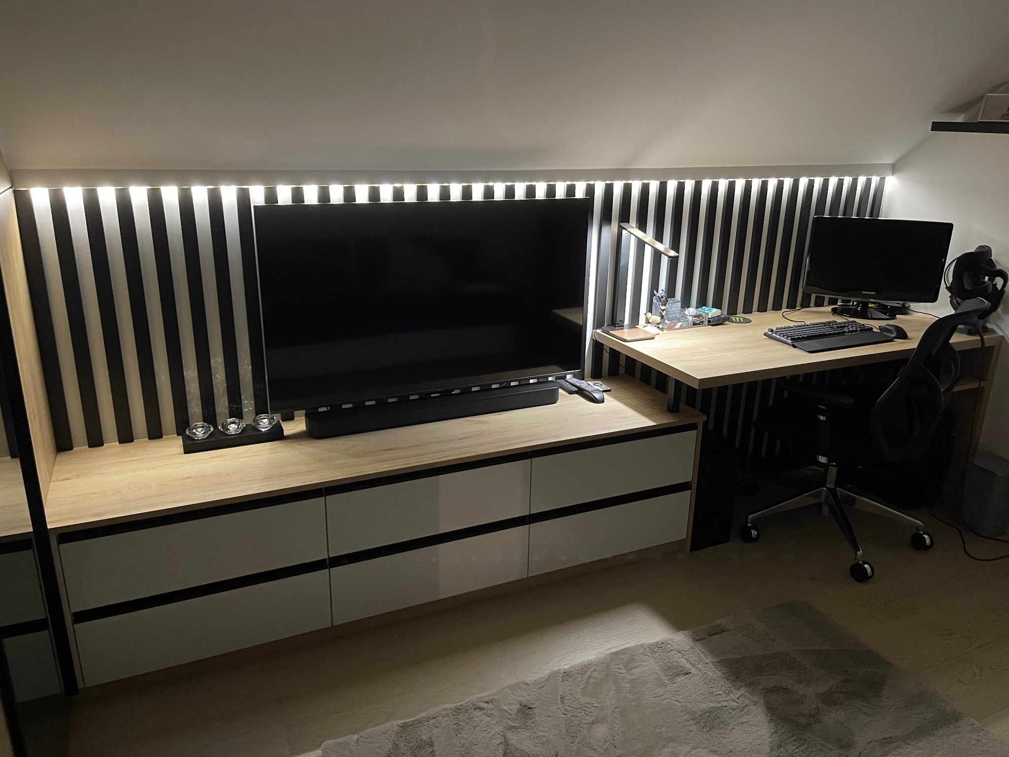 Zabudowa pod skosem w Jastrzębiu-Zdroju z szafkami, blatem i czarno-białymi panelami ściennymi, tworząca kącik telewizyjny i miejsce do pracy z biurkiem i komputerem.