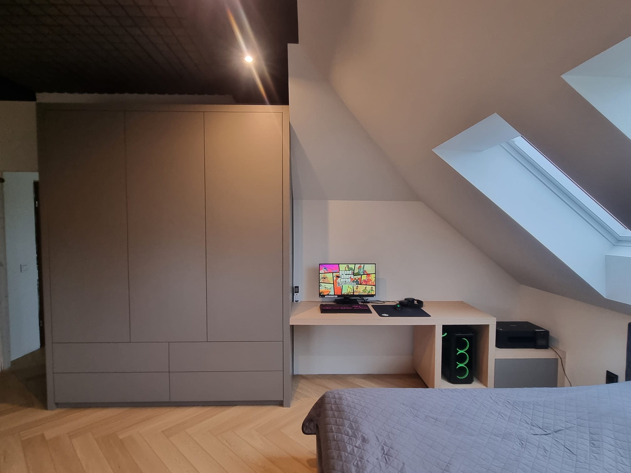 Szara szafa wnękowa z biurkiem w pokoju na poddaszu, widoczny komputer stacjonarny z podświetleniem LED i gra na ekranie.