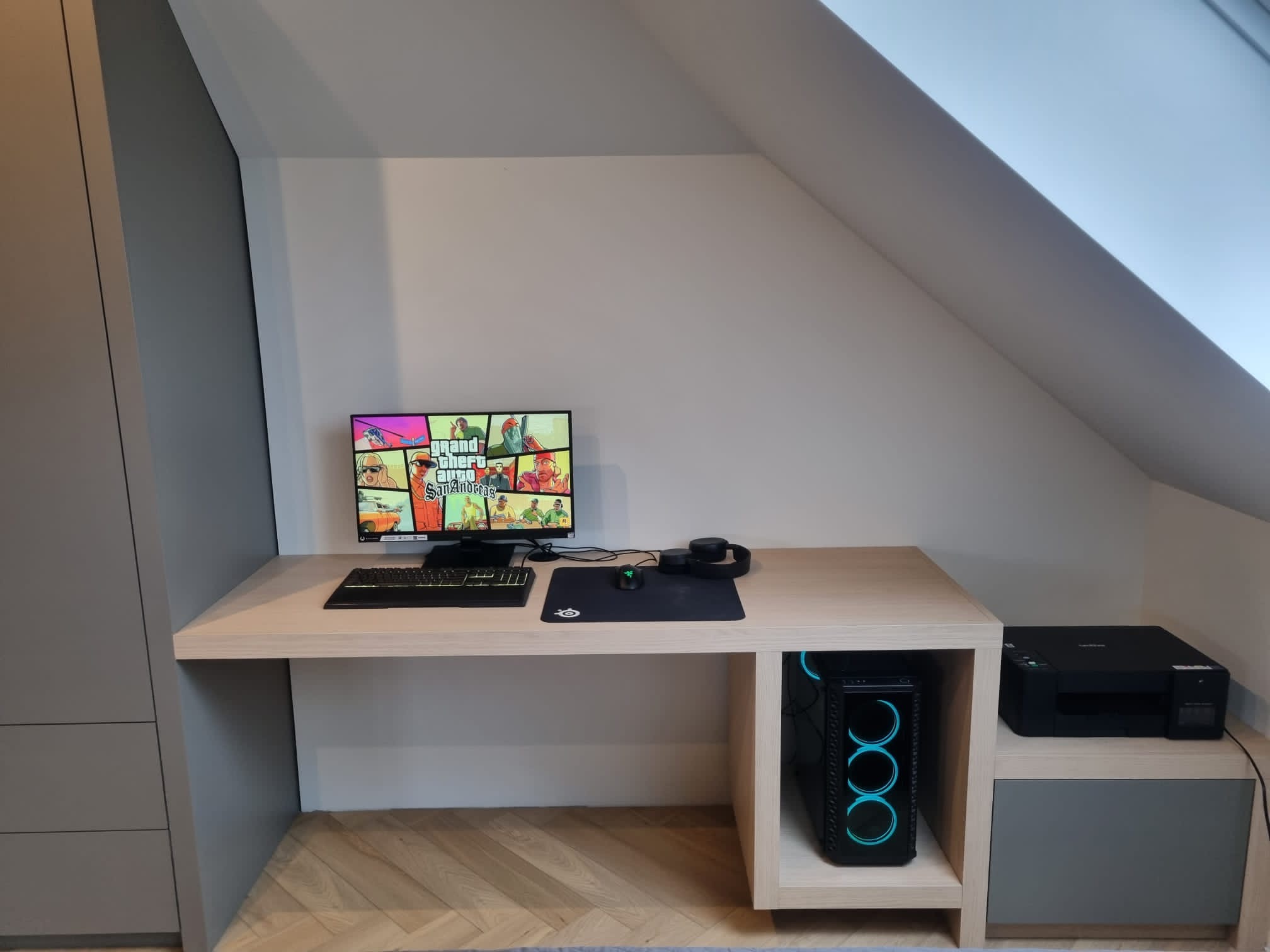 Komputer stacjonarny z podświetleniem LED umieszczony w przestrzeni biurka na wymiar, monitor wyświetla grę Grand Theft Auto, obok drukarka, całość w pokoju na poddaszu.