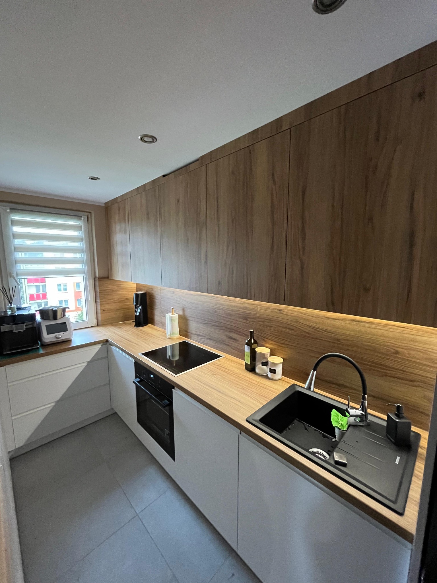 Nowoczesna kuchnia z drewnianymi szafkami górnymi i białymi dolnymi, blatem imitującym drewno oraz czarnym zlewem z baterią i dozownikiem na mydło. Na blacie stoją przyprawy i butelka z oliwą.