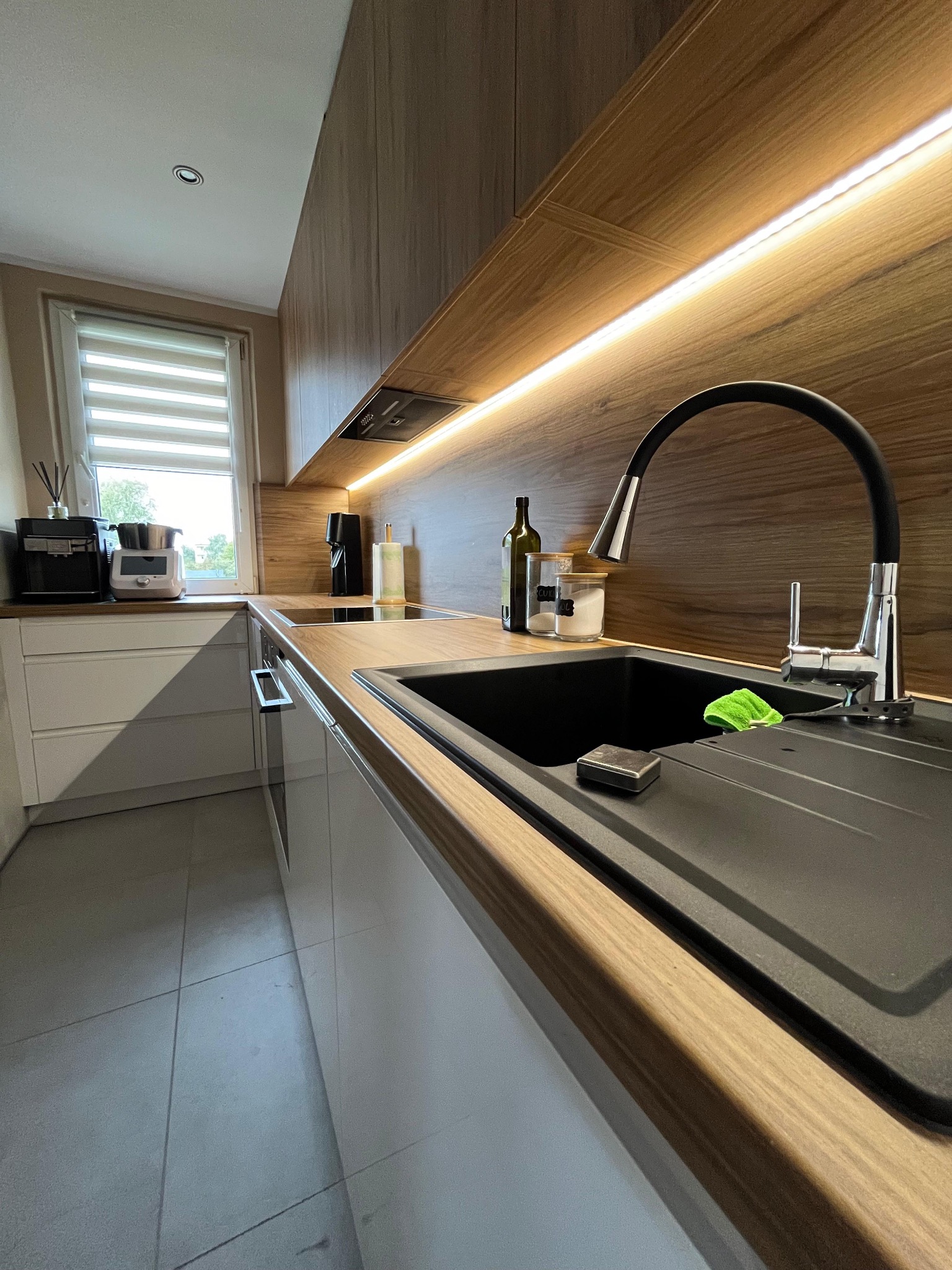 Nowoczesna kuchnia z blatem drewnianym, czarnym zlewem i baterią z elastyczną wylewką. Podświetlenie LED pod szafkami górnymi. Na blacie butelka oliwy i pojemniki na przyprawy.