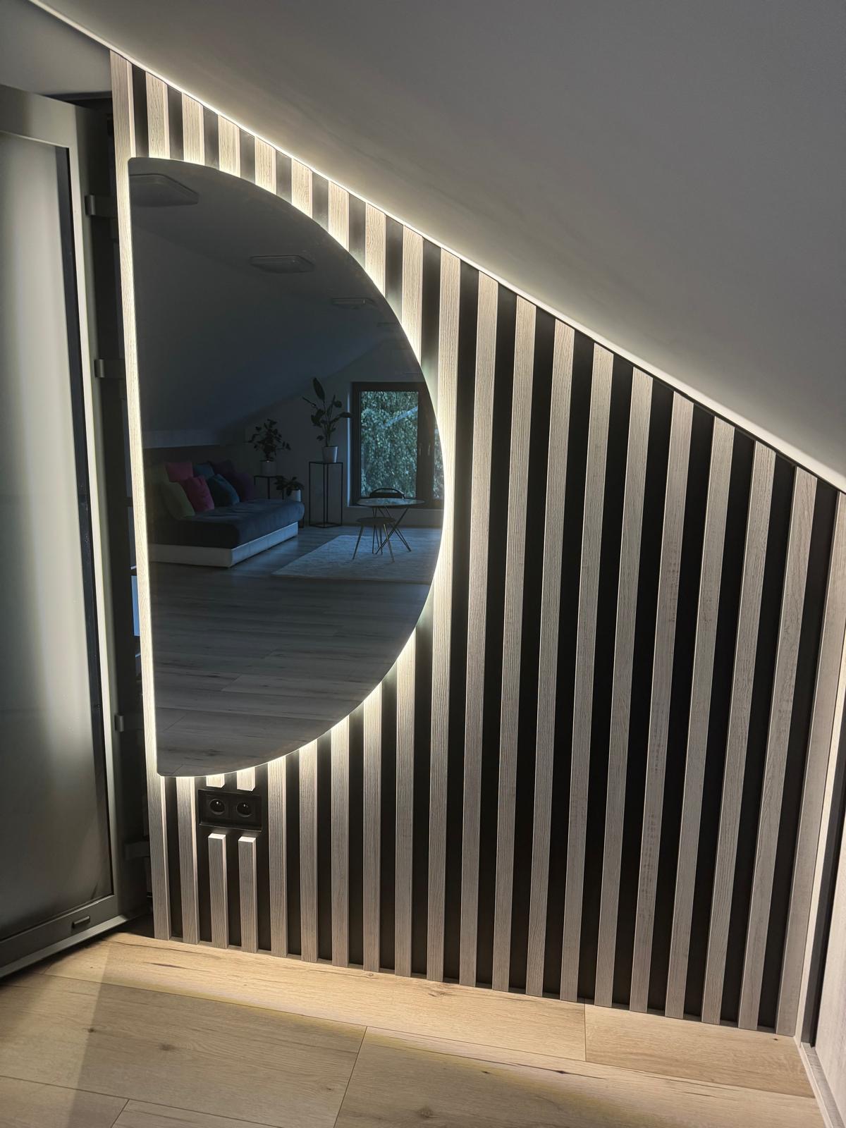 Ściana z drewnianych lameli z wbudowanym, podświetlanym lustrem w kształcie półksiężyca, odbijającym wnętrze pokoju na poddaszu z szarą sofą i roślinami.