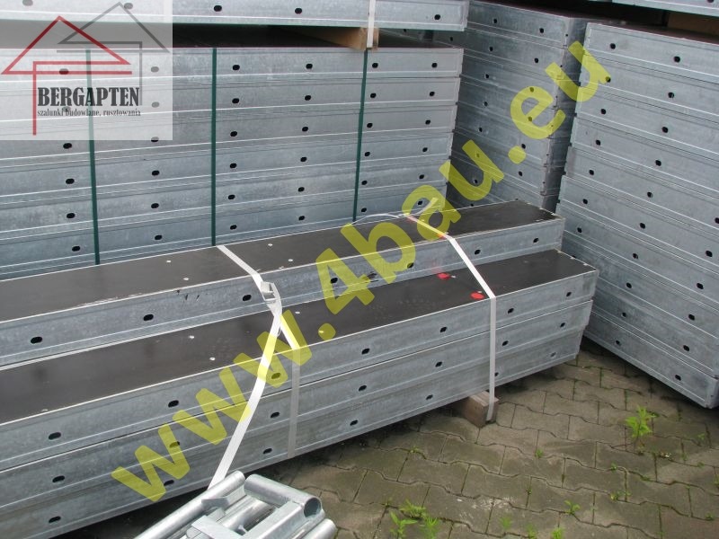 Sztaple szalunków budowlanych z metalową ramą i czarnym wypełnieniem, zabezpieczone taśmą, składowane na zewnątrz. Widoczne otwory montażowe.