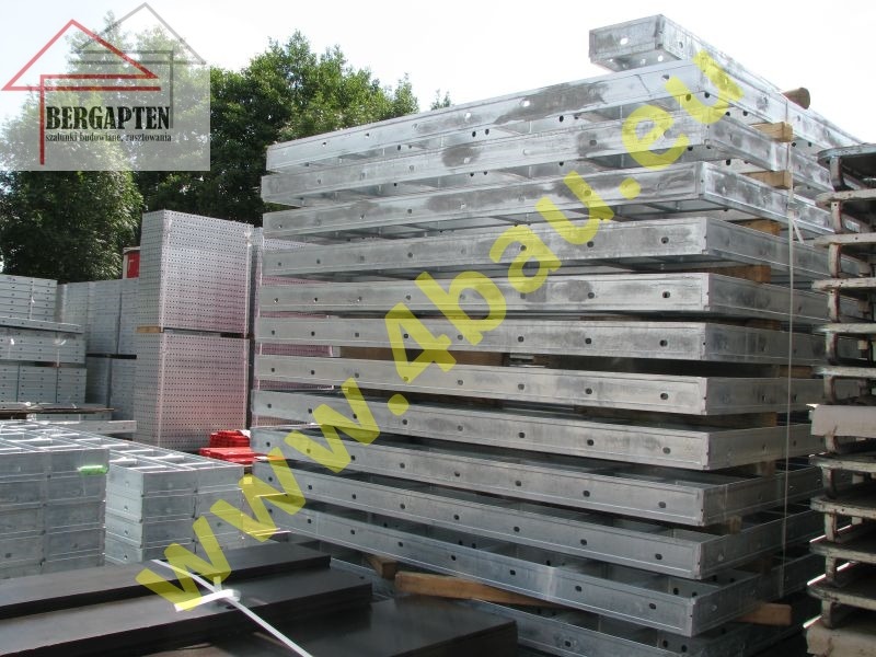 Stos szalunków budowlanych z metalu, ułożonych na palecie. Widoczne otwory montażowe i logo producenta w lewym górnym rogu. Tło stanowią drzewa.