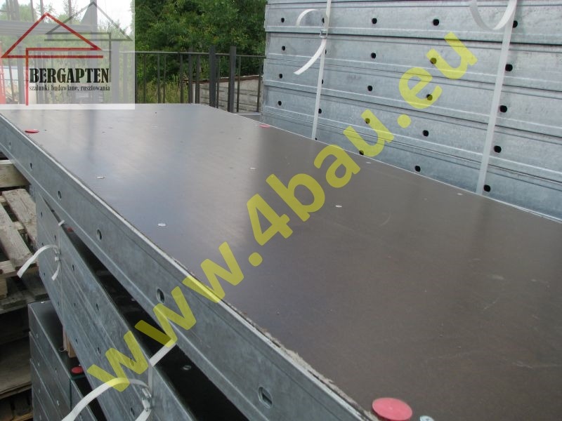 Stos płyt szalunkowych z metalową ramą, zabezpieczonych taśmami. Widoczne otwory w ramie i czerwone zaślepki. Tło z zielenią.