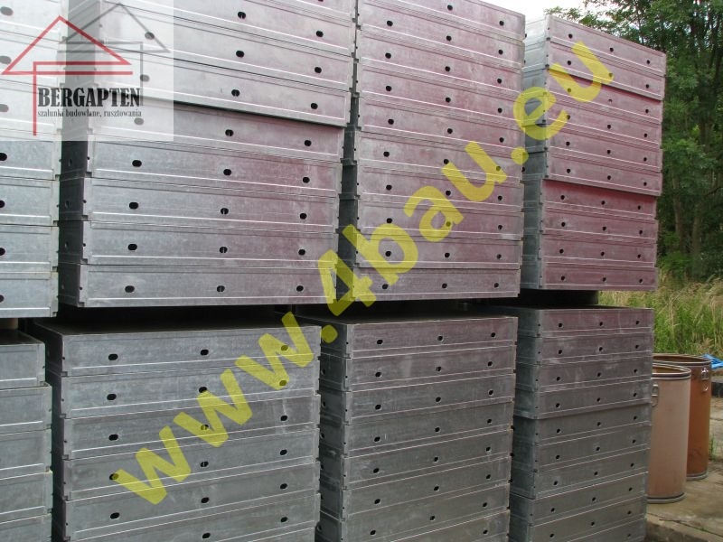 Stos szalunków budowlanych z otworami, ułożonych jeden na drugim. Szalunki w kolorze srebrnym, widoczne elementy konstrukcyjne i tło z zielenią.