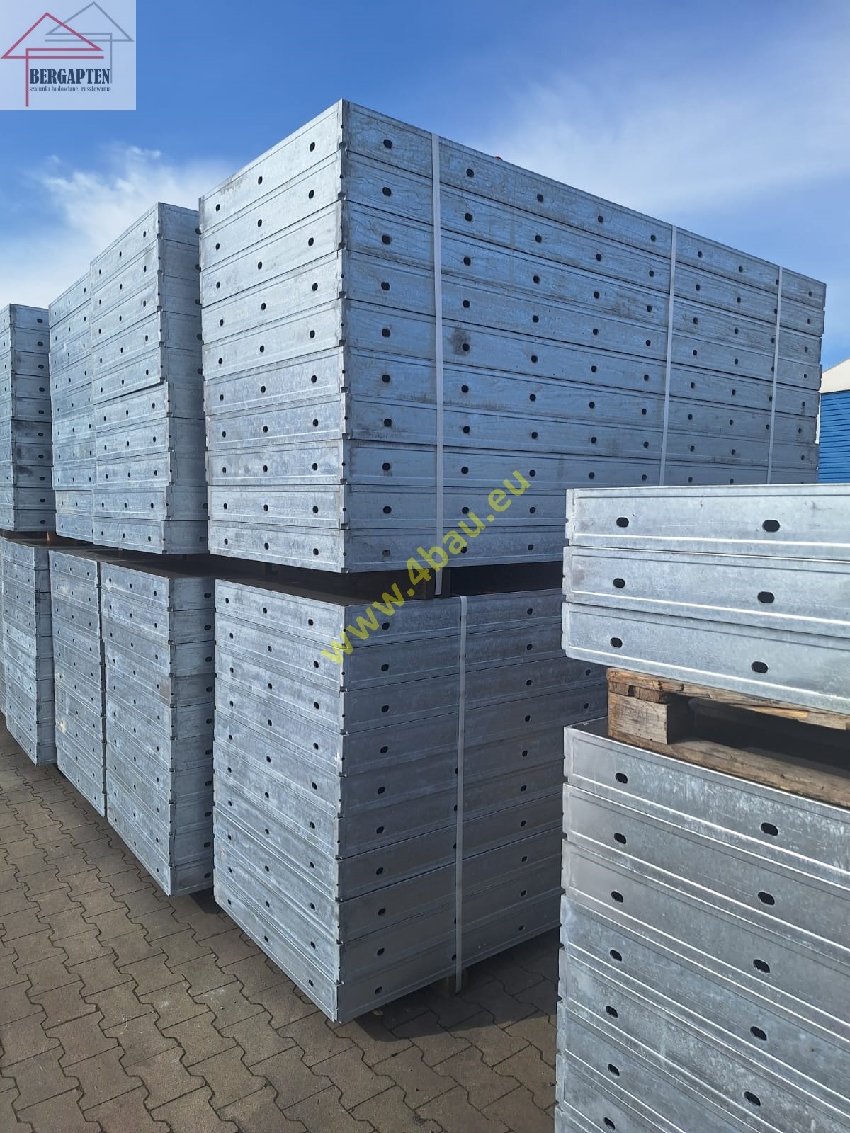 Stosy szalunków budowlanych z metalu, spięte taśmami, ułożone na paletach. Widoczne otwory montażowe. Tło: błękitne niebo i fragment niebieskiego budynku.