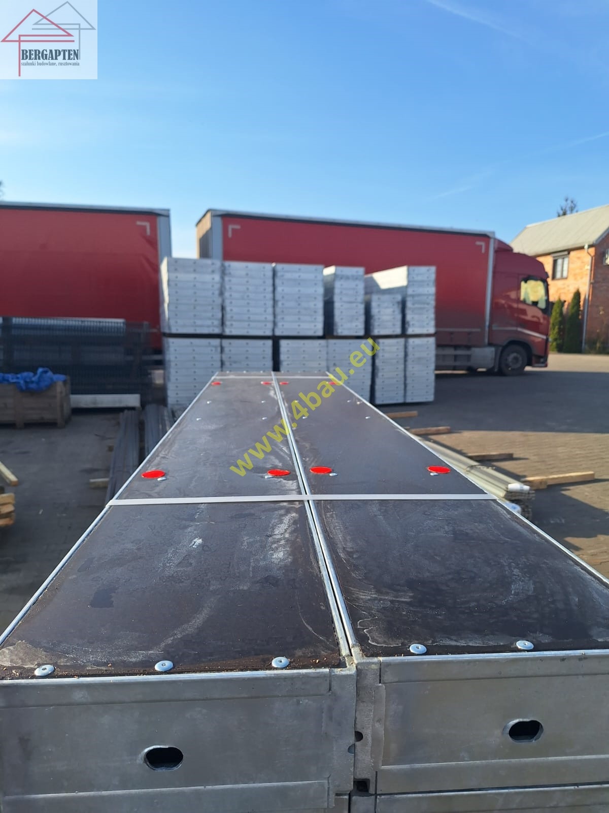 Stos paneli szalunkowych z czarną powierzchnią i czerwonymi zaślepkami, na tle czerwonych ciężarówek i błękitnego nieba. Widok z bliska, z perspektywy.