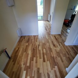 Panele Winylowe-Laminowane - Jasna podłoga z drewnianych desek ułożona w korytarzu. Widoczne przejście do pokoju z oknem. Na ścianie biała skrzynka. U dołu fragment schodów.