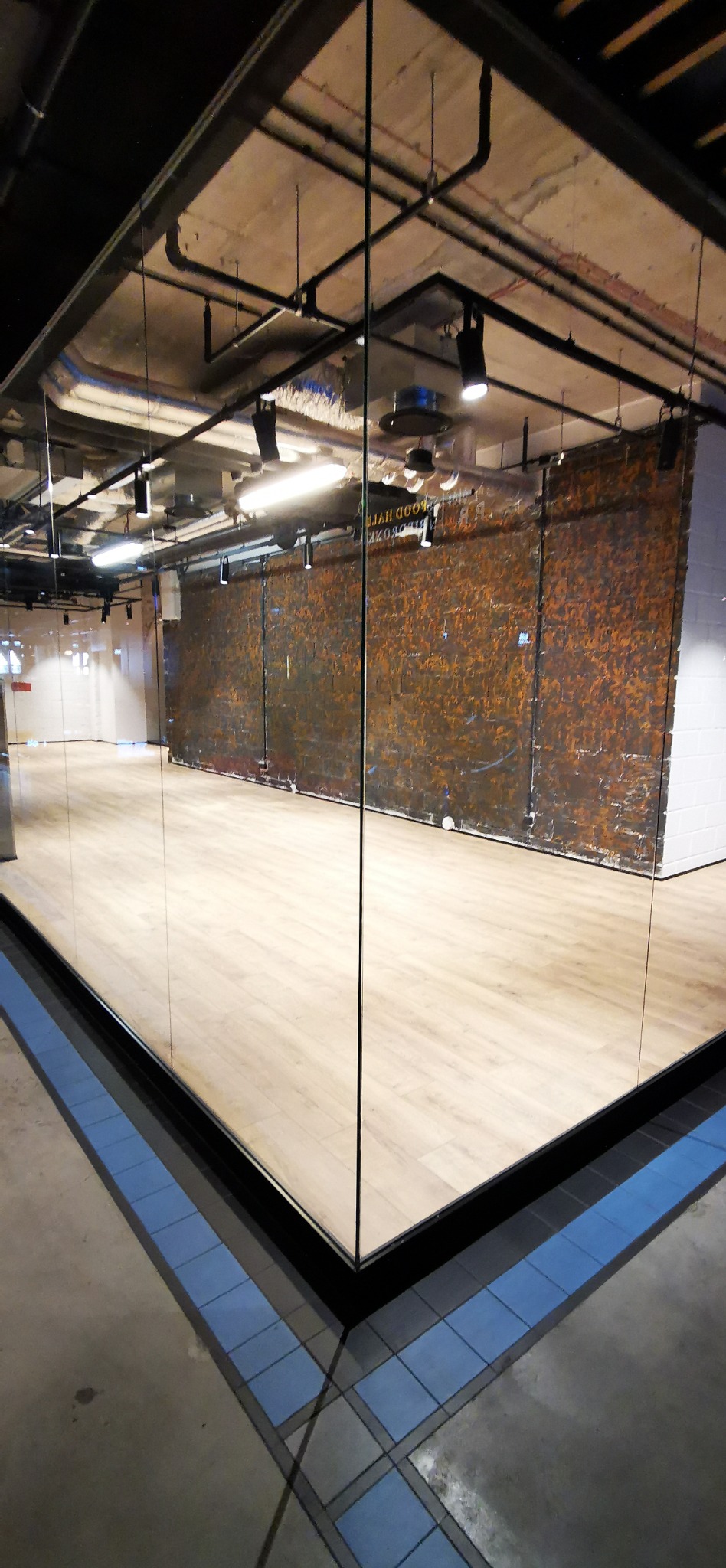 Widok na salę z jasnymi panelami podłogowymi, widoczne instalacje pod sufitem i ściana z cegły. Przezroczysta ściana oddziela przestrzeń od korytarza z niebieską mozaiką.