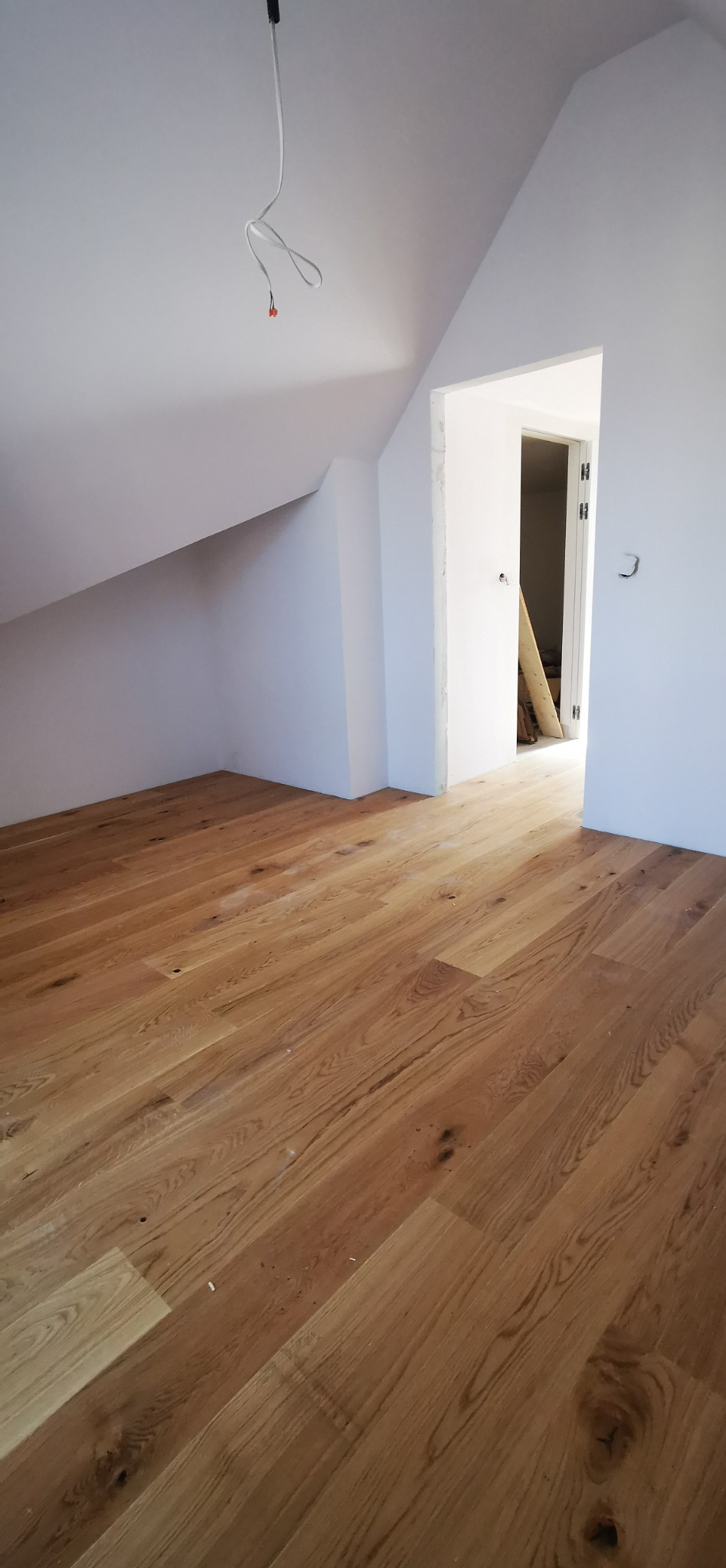 Jasna, drewniana podłoga z paneli w nowym pokoju na poddaszu, widoczne kable zwisające z sufitu i surowy otwór drzwiowy.