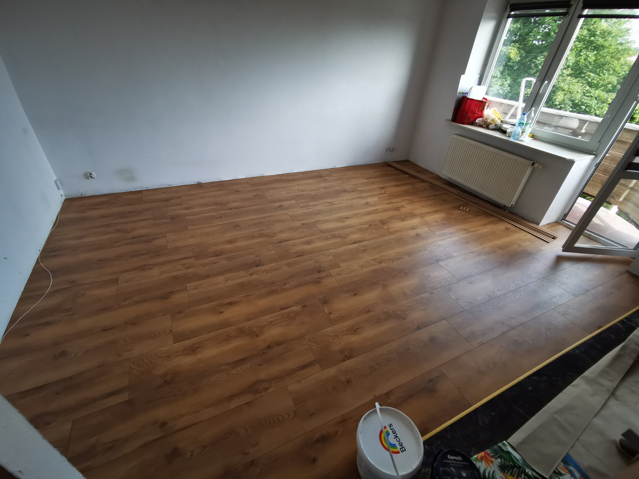 Układanie paneli podłogowych w pokoju, widoczny fragment ułożonej podłogi z paneli w odcieniach brązu, białe ściany, okno z widokiem na zieleń, resztki paneli przy ścianie, wiadro z farbą Beckers...