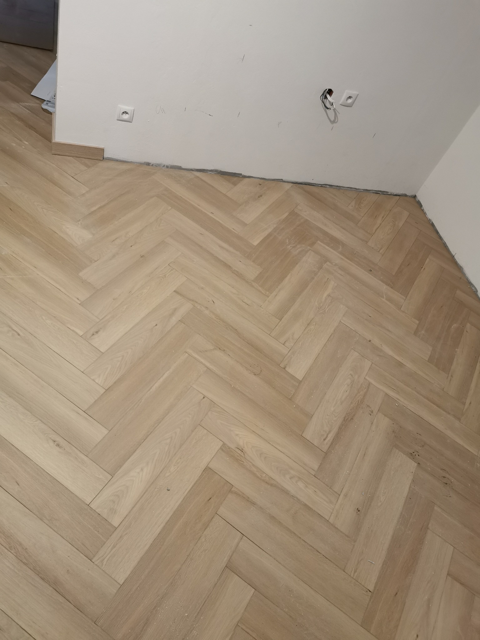 Parkiet ułożony w jodełkę francuską, widoczne gniazdka elektryczne i niedokończone listwy przypodłogowe.