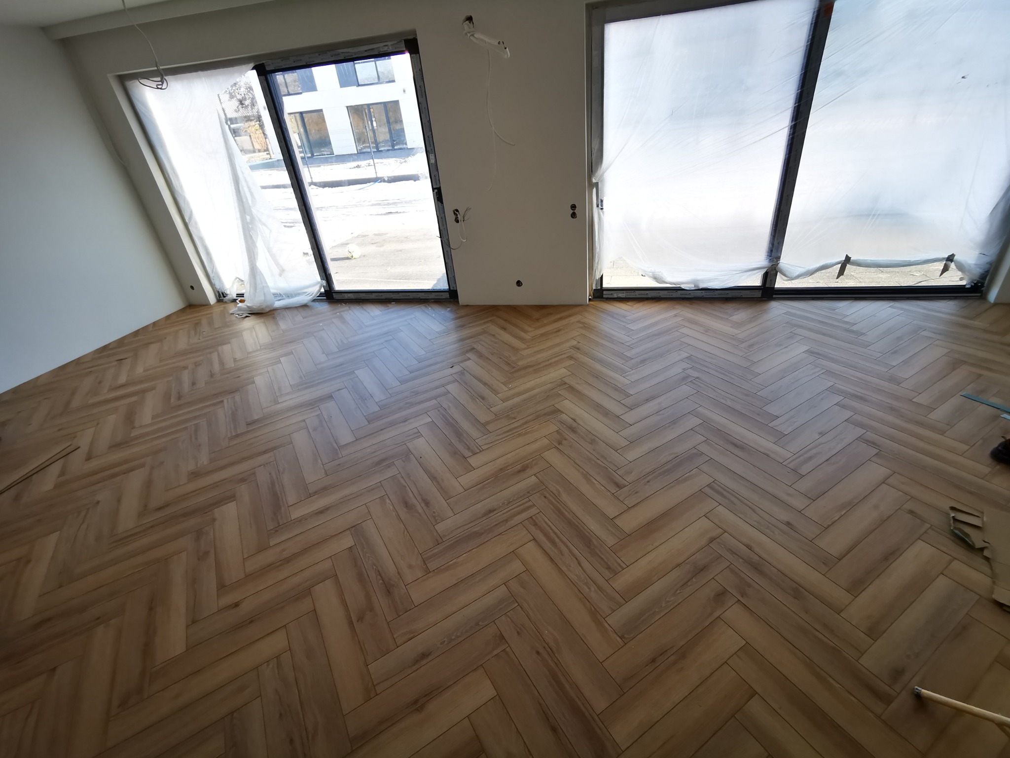 Ułożona podłoga z paneli w jodełkę, widok na okna zabezpieczone folią malarską. Jasne panele, białe ściany, surowy stan wykończenia wnętrza.